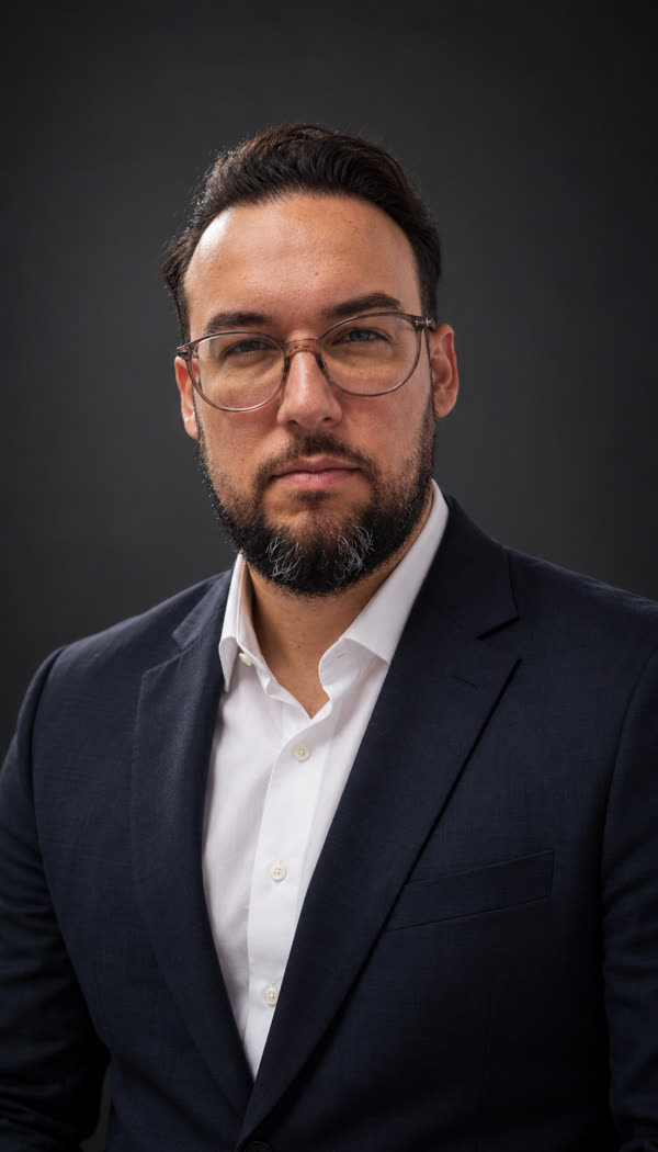 Bruno Lima, de óculos, camisa branca e blazer azul-marinho, sobre fundo escuro